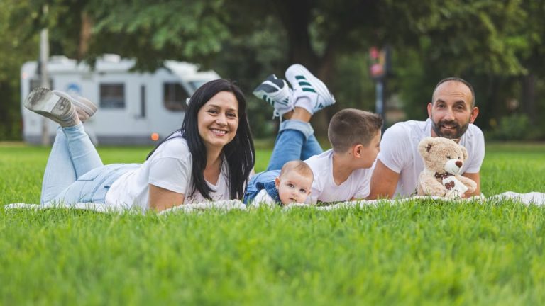 Happy family lying on grass with RV in the background, enjoying a sunny outdoor picnic.